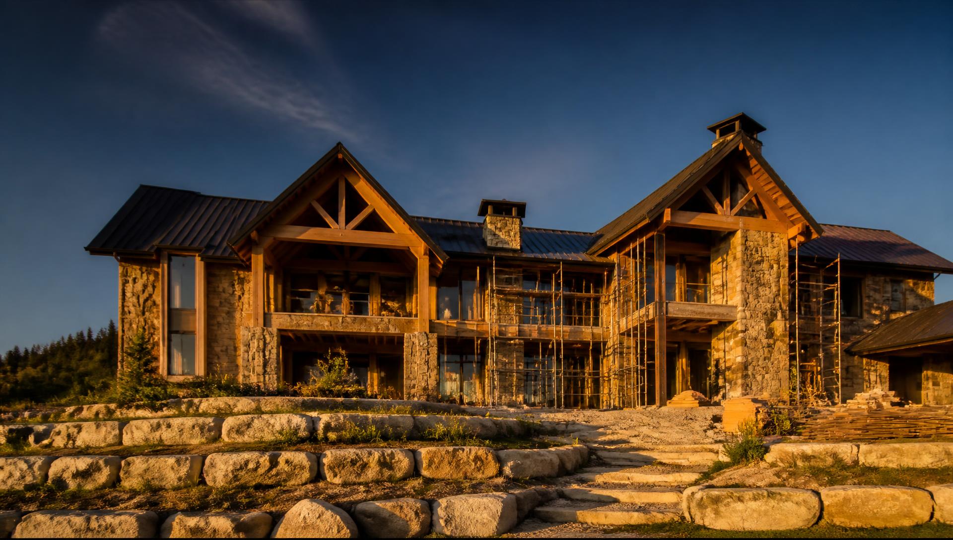 A large custom home under construction at golden hour with timber framing, stone facade, and scaffolding in place